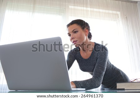 Young attractive professional business woman at her office space work desk reaching and typing on her laptop computer against a bright window, working. Business finance and working on a job, interior.