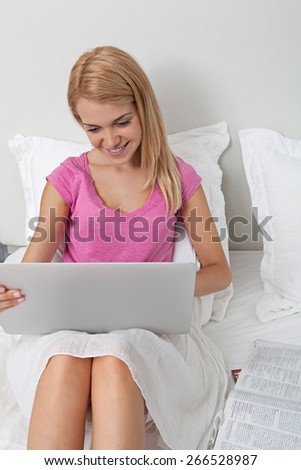 Beautiful teenager woman relaxing on a bed in a home bedroom using a laptop computer and magazines, smiling house interior. Connectivity technology buying and spending lifestyle, hotel room interior.