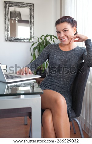Portrait of an attractive young office business woman sitting joyfully smiling in her home office desk, workplace interior. Professional working woman using technology at work, indoors.