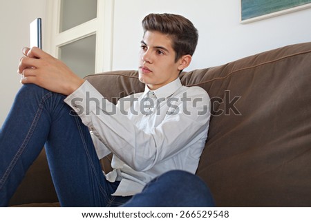 Side portrait of a fashionable teenager young man wearing a shirt lounging on a sofa at home, using a smart phone device for networking on line, home interior. Young modern man lifestyle indoors.