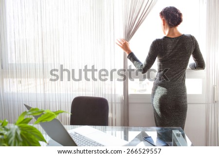 Rear view of a office business woman standing looking out a bright window in her home office desk, thoughtful and contemplative, interior. Aspirational professional woman using technology, indoors.