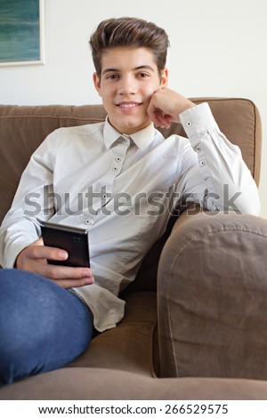 Portrait of a fashionable teenager young man wearing a shirt and lounging on a sofa at home, using a smart phone for networking on line, home interior. Young modern man smiling, lifestyle indoors.