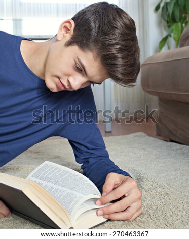 Portrait of attractive teenager man holding a text book open while laying down on a rug in a home living room with a sofa, reading and studying at home, interior. Student living, interior lifestyle.