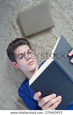 Close up portrait of a young student man reading a book laying down on a bed at home. Boy studying at home laying on a carpet doing his homework and learning indoors. Student home life.