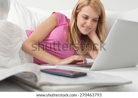 Portrait of attractive woman relaxing in a stylish decorative white home bedroom using a laptop computer, with magazines and a smartphone smiling, interior. Home technology lifestyle, indoors.