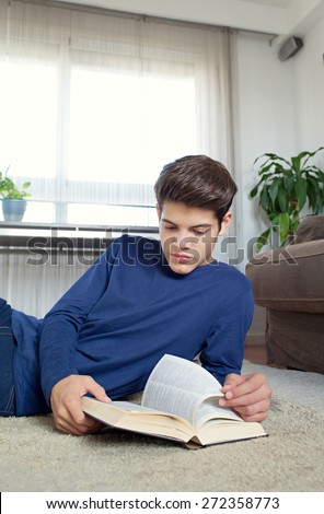 Portrait of a student teenager man holding a text book open laying on rug in home living room, reading and studying at home, interior. Student boy doing homework in house living, interior lifestyle.