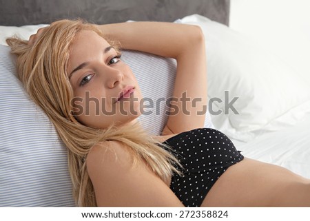 Beauty portrait of young woman relaxing on a luxury bed in a home bedroom, thoughtfully looking at camera wearing sexy black bra lingerie, hotel room, indoors. Home living lifestyle care, interior.