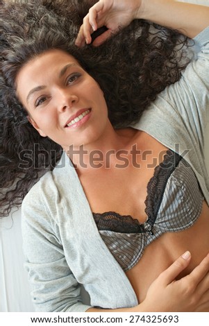 Overhead portrait of a beautiful exotic woman relaxing on a bed at home, smiling wearing sexy lingerie, interior space. Healthy wellness and well being living aspirational lifestyle, bedroom indoors.