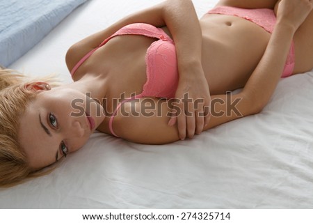 Overhead beauty portrait of young woman laying on a luxury bed in a home bedroom, smiling flirting wearing sexy pink bra lingerie, hotel room, indoors. Home lifestyle and skin hair care, interior.