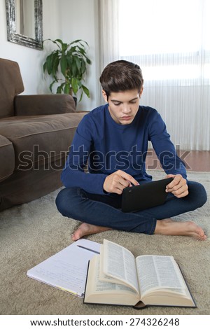 Thoughtful teenager boy sitting on a rug in a house living room using a digital tablet pad to do his homework at home. Young student man using telecommunications technology, home living lifestyle.