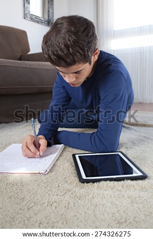 Teenager boy on a rug in a house living room using a digital tablet pad to do his homework and study at home. Young student man learning using telecommunications technology, home living lifestyle.