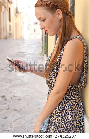 Side view of a young woman sightseeing visiting a picturesque stone pavement street, using a smartphone mobile, holding a map in a destination city on summer holiday, outdoors. Travel technology.