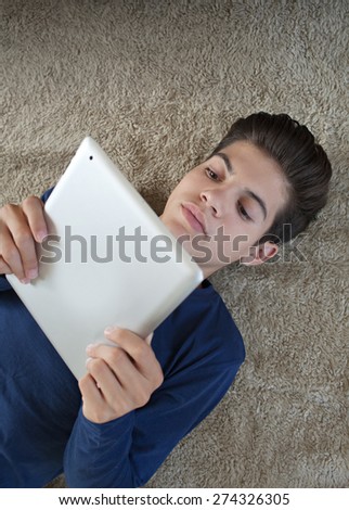 Over head portrait of a teenager boy laying on a rug in a house living room using a digital tablet pad to study at home. Young student man using telecommunications technology, home living lifestyle.