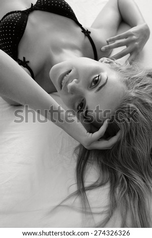 Black and white beauty portrait of young woman laying on a luxury bed in a home bedroom, smiling flirting, wearing sexy black bra lingerie, indoors. Home lifestyle and skin hair care, interior.