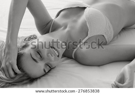 Black and white close up beauty portrait of young woman relaxing on a bed in a home bedroom, smiling flirting, wearing sexy bra lingerie, hotel room, indoors. Home lifestyle skin hair care, interior.