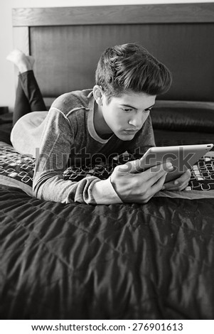 Black and white portrait of young teenager man laying on a bed at home, relaxing and using a digital tablet, interior. Lifestyle technology at home. Young person using technology indoors, thoughtful.