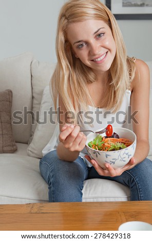 Portrait of attractive young woman sitting on sofa in a home family room living room, eating a fresh colorful salad, smiling. Girl eating healthy food at home, interior. Well being lifestyle.