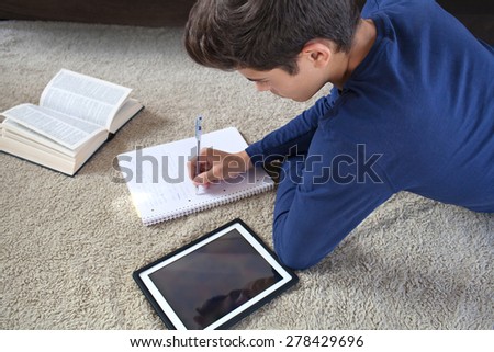 Rear side view of a teenager boy laying on a rug in a house living room using a digital tablet to study at home. Young student man learning using telecommunications technology, home living lifestyle.