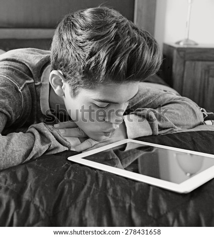 Black and white portrait of young teenager man laying on his bed at home, relaxing using a digital tablet, interior. Lifestyle technology at home. Young person using technology indoors, thoughtful.