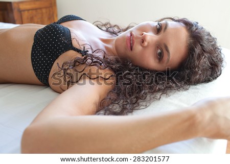 Portrait of beautiful young exotic woman laying on a bed at home relaxing and being sexy wearing sensual black lingerie, looking at camera with a moody expression. Aspirational lifestyle, interior.