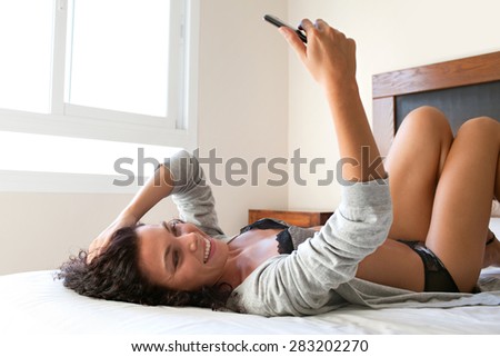 Side view of a beautiful young woman laying on a bed at home, holding up and using a smartphone device to take selfies pictures of herself, networking on line, home interior lifestyle.