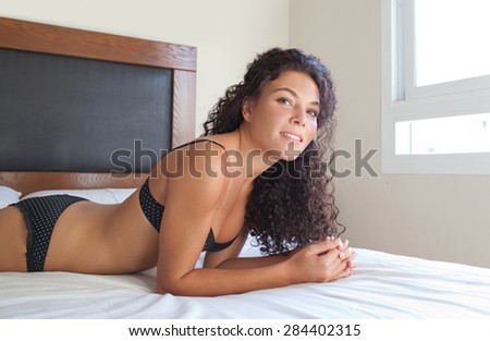 Beauty body portrait of young beautiful exotic sexy woman laying on a bed at home in black lingerie, smiling and looking at the camera relaxing in a bedroom, interior. Aspirational lifestyle, indoors.