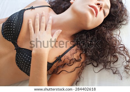 Portrait of beautiful young exotic woman laying down on a bed at home relaxing and being sexy wearing sensual black lingerie bra with her eyes closed. Aspirational lifestyle and well being, interior.