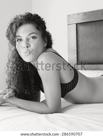 Black and white sexy body of young woman laying on a bed at home in black lingerie, sensual, playful and blowing a kiss to the camera, happy. Beauty, well being and care in a home bedroom, interior.