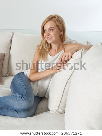 Beautiful young blond woman sitting and relaxing on a white sofa at home, smiling and lounging in a stylish and elegant living room, luxury spacious home interior. Aspirational lifestyle, indoors.