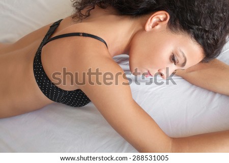 Beauty body view of a young beautiful exotic sexy woman laying on a bed at home in black lingerie, being thoughtful and moody, relaxing in a bedroom, interior. Health well being lifestyle, indoors.