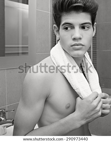 Beauty portrait of a young man in a home bathroom with a bare chest and a white towel around his neck, thoughtful, home interior. Health, care, male grooming, indoors. Man portrait with perfect skin.