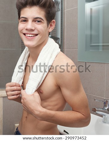 Beauty portrait of a young man in a home bathroom with a white towel around his neck, joyfully laughing and looking at camera, interior. Health male grooming, indoors. Man portrait with perfect skin.