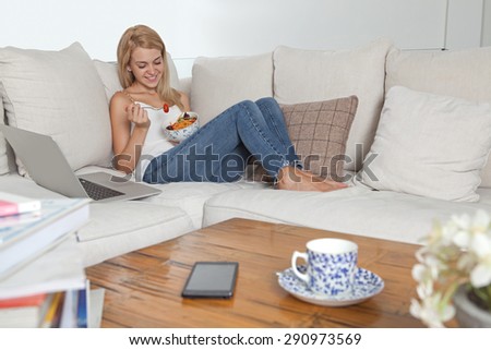Beautiful young blond woman sitting on a white sofa at home, eating a healthy salad in living room, using a laptop computer, interior. Home technology lifestyle and healthy eating habits, indoors.