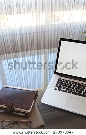 Still life view of an office room with an open laptop computer on a glass desk with reflections against a window, office interior with paperwork. Slick and professional workplace, technology indoors.