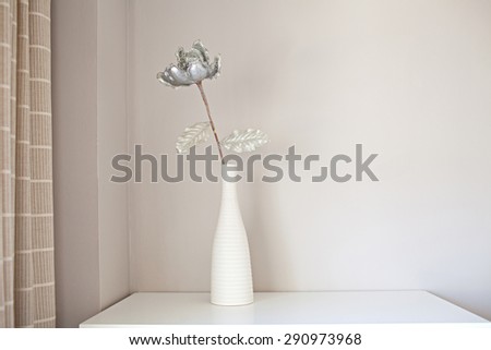 Still life decorative view of a fake silver glitter flower in a white vase in a home living room, against a plain wall with curtain, home interior. Home decoration detail, indoors with wall space.