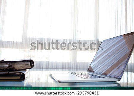 Still life of an office room with an open laptop computer on a glass desk with bright reflections against a window, office interior with paperwork. Slick professional workplace, technology indoors.