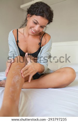 Body view of a beautiful young woman applying cream with her hands on her legs skin on a white bed at home smiling, wearing a robe and lingerie, bedroom interior. Health, wellness and care, interior.