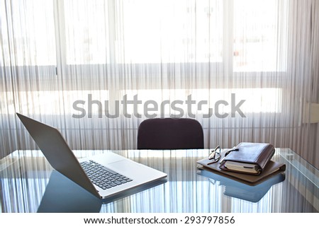 Still life of office interview room with an open laptop computer on a glass desk with reflections against window, office interior with paperwork. Slick and professional workplace, technology indoors.
