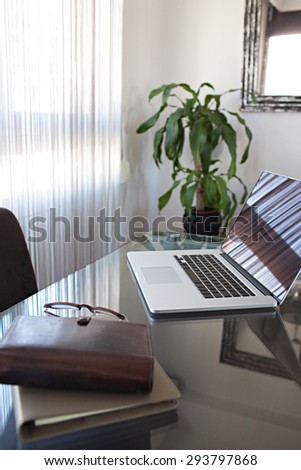 Still life of an office room with an open laptop computer on a desk with reflections against a window, office interior with paperwork and chair. Slick and professional workplace, technology indoors.