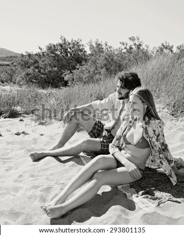 Black and white view of attractive young couple relaxing together, sunbathing on beach on summer holiday, enjoying nature outdoors. Travel lifestyle and recreational living, exterior with mountains.