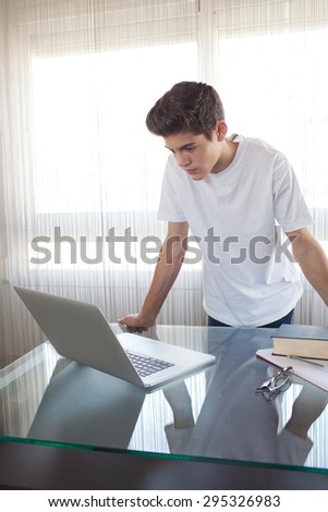 Teenager boy standing at a desk at home doing his homework using a laptop computer and books, being thoughtful, interior. Adolescent using technology, studying by bright window, indoors.