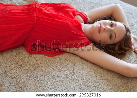 Over head beauty portrait of an attractive young woman laying on a bed, in a bright red dress, looking happy and thoughtful at the camera, home interior. Beautiful woman lifestyle, relaxing indoors.