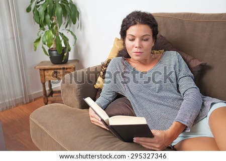 Portrait of beautiful young woman lounging on a comfortable sofa at home, reading a book on holiday, living room interior. Relaxing home living lifestyle, indoors. Girl reading a book on the weekend.
