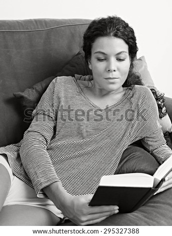 Black and white portrait of beautiful young woman lounging on a sofa at home, reading a book while on holiday, living room interior. Relaxing home living lifestyle, indoors. Girl reading thoughtful.