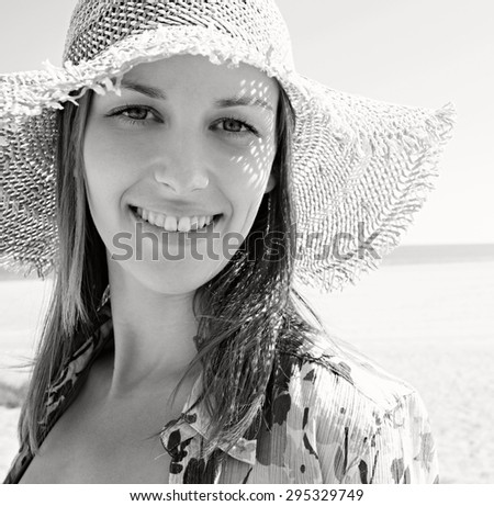 Black and white beauty portrait of a young tourist woman on a spacious sand beach with mountains, smiling and in a straw hat during a sunny summer holiday, outdoors. Travel recreation lifestyle.
