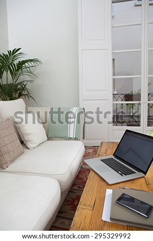 Still life detail view of a living room with sofa, an open laptop computer with blank screen and a smartphone with paperwork and folders, home interior. Aspirational technology indoors lifestyle.