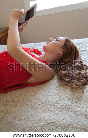 Side portrait of a beautiful young woman holding and using a digital tablet laying down on a bed at home, interior. Girl using technology, laying down relaxing and lounging in bedroom, indoors.