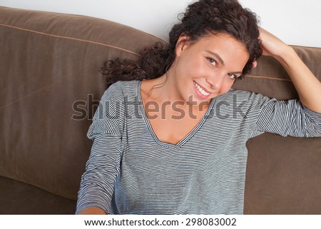 Beautiful and joyful young woman lounging on a comfortable sofa at home, relaxing in living room interior. Positive home lifestyle, indoors. Girl calm and tranquil leaning on hand, smiling.