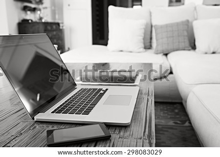Still life black and white detail of luxury living room with sofa, open laptop computer and smartphone with paperwork and folders, home interior. Aspirational technology living, indoors lifestyle.