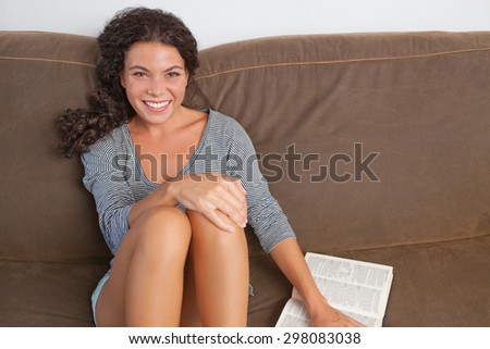 Portrait of beautiful young joyful woman lounging on a comfortable sofa at home reading a book, relaxing in living room interior. Enjoying fun home lifestyle, indoors. Girl reading weekend book.